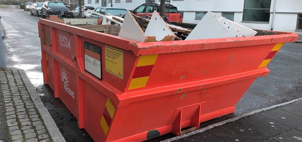 En orange container full med skräp o gamla möbler står på en gata med parkerade bilar.